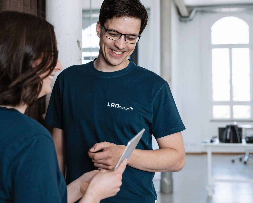 Zwei junge und fröhliche Menschen stehen im Flur, die Dame im Vordergrund hält ein Tablet in der Hand und zeigt ihrem Kollegen die neue Cloudsoftware (SaaS) LANcloud und deren Vorteile.
